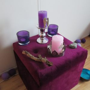 Auf einem gepolsterten Meditationsaltar befinden sich Kerzen in rosa und lila sowie ein Ast und ein Stein.